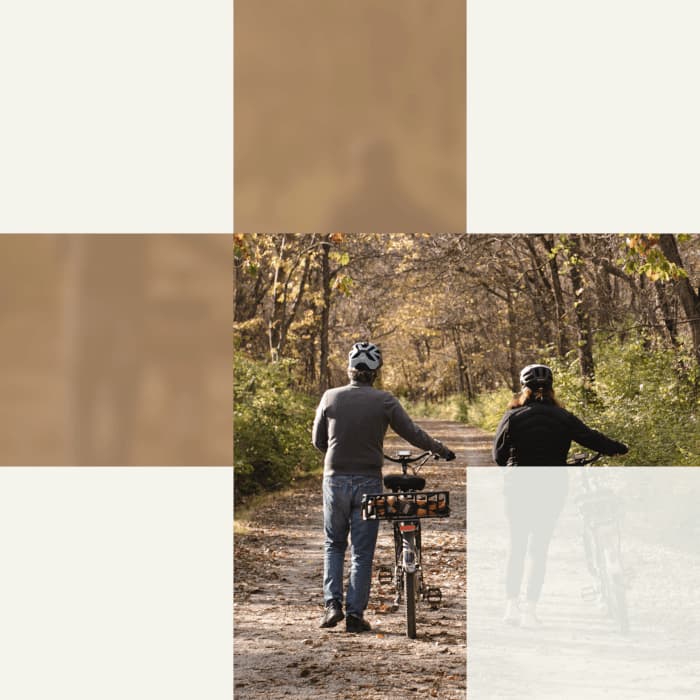 Two people walking on a dirt path in a wooded area, each holding a bicycle. One person is wearing a helmet and a gray sweater, while the other is wearing a black jacket and helmet. The scene is set in autumn with leaves on the ground.