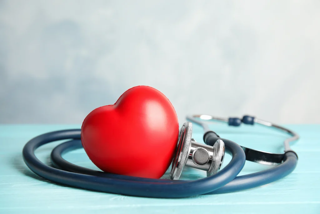 A red heart-shaped object resting next to a stethoscope on a light blue wooden surface.