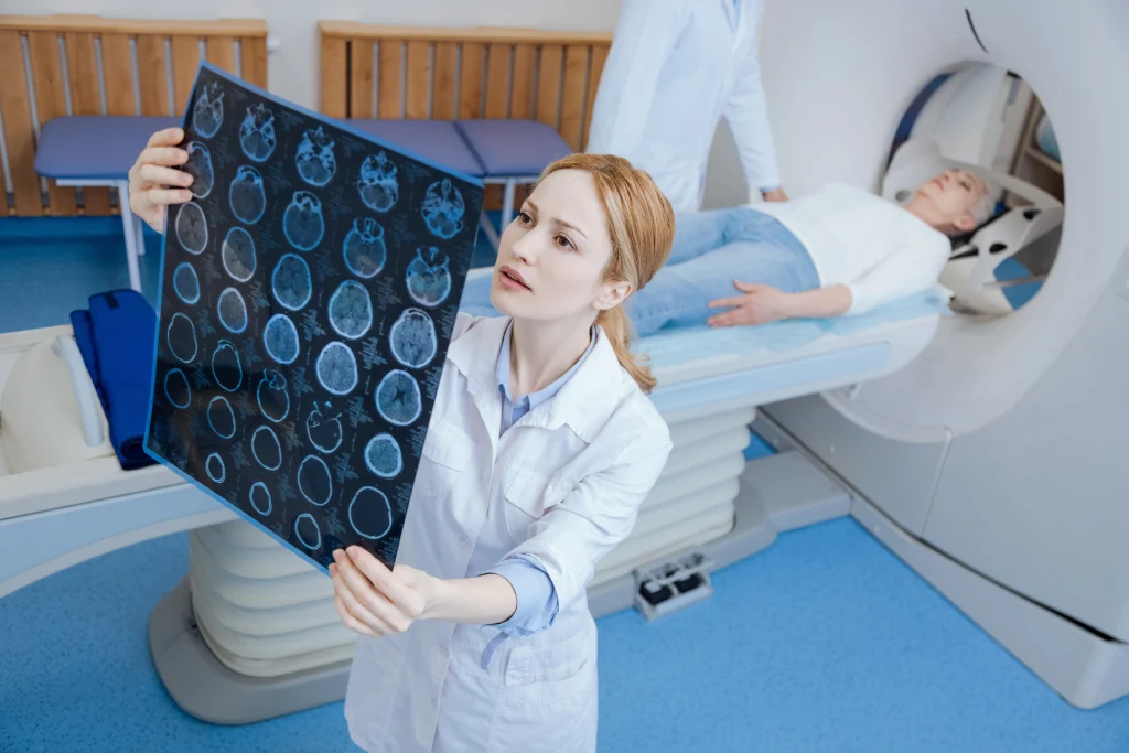 A medical professional in a lab coat holds up a brain scan while examining it, with a patient lying on a medical imaging table in the background. Another medical staff member is positioned near the imaging machine.
