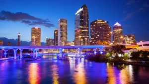 A vibrant city skyline at dusk, featuring illuminated skyscrapers and a reflective river. The scene includes a bridge with blue lighting, palm trees, and a city with various architectural styles under a deep blue sky.