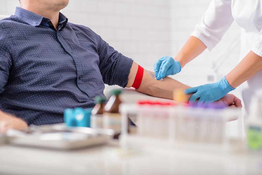 A healthcare professional in blue gloves prepares to draw blood from a patient wearing a navy shirt with a red tourniquet on their arm. In the background, there are medical supplies, including bottles and test tubes.