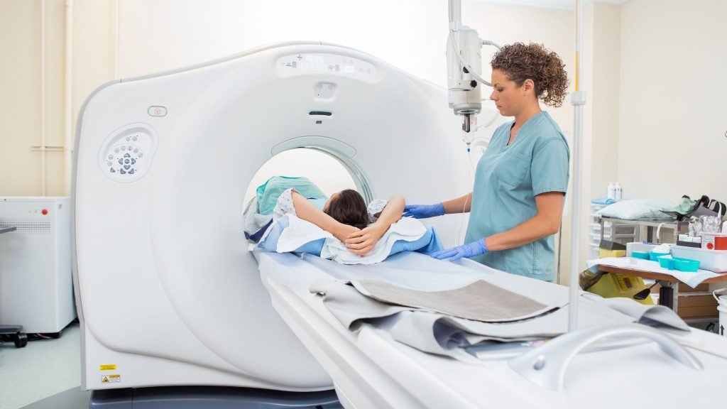 A medical technician assists a patient lying on a CT scan machine, preparing for the scan. The patient is wearing a hospital gown and has their arms positioned behind their head, while the technician is dressed in scrubs and wearing gloves. Medical equipment and supplies are visible in the background.
