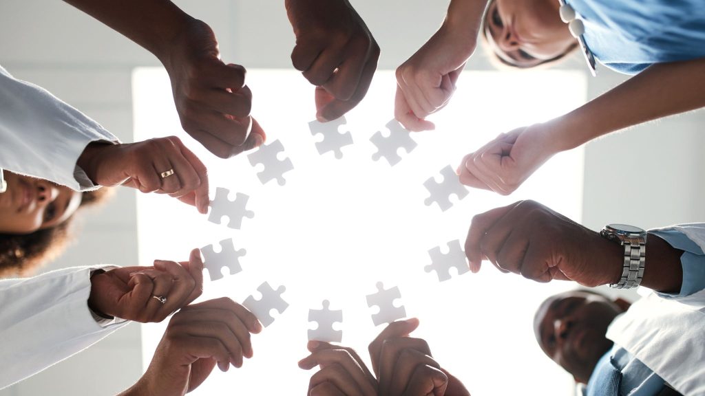 A group of diverse hands reaching toward the center, each holding white puzzle pieces against a bright light source in the background.