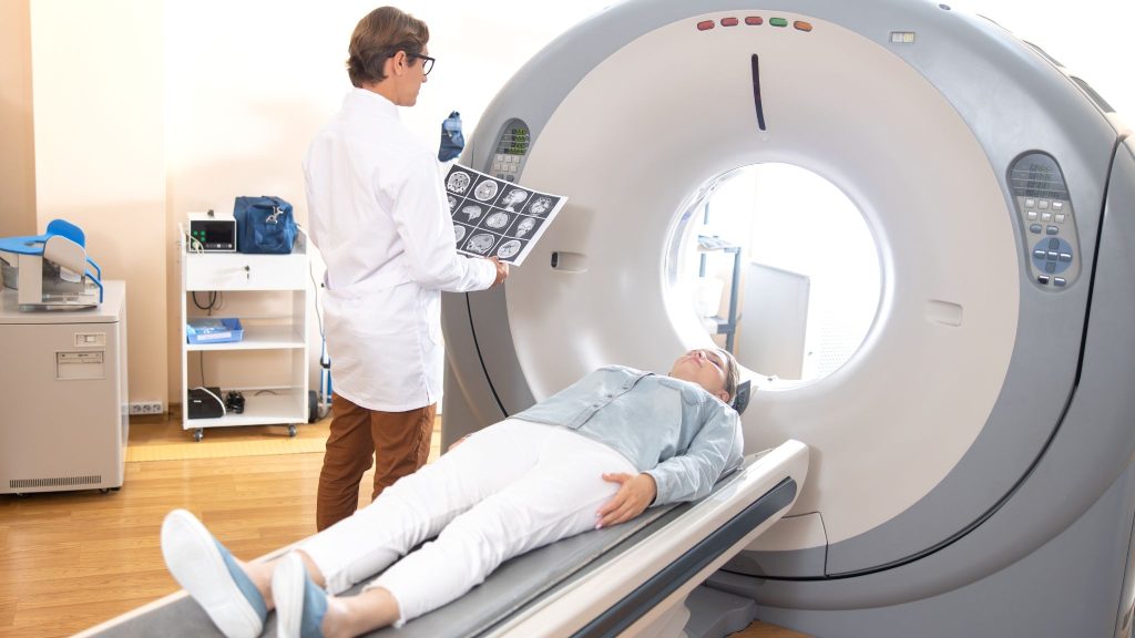 A doctor in a white coat holds medical imaging results while a patient lies on a CT scan machine, preparing for a scan.