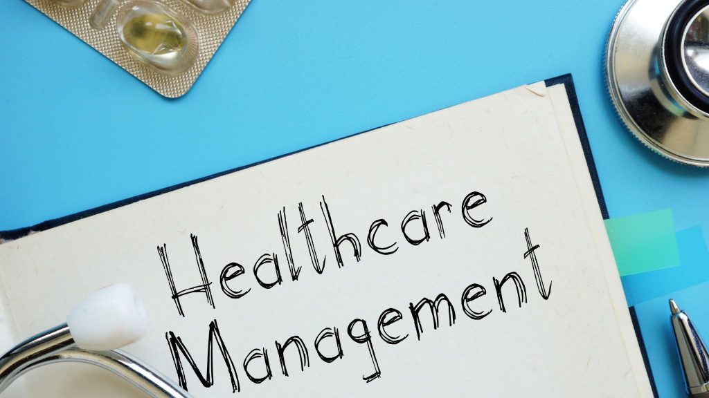 A stethoscope, a pen, and a notebook with the text "Healthcare Management" prominently displayed on a blue background, alongside a blister pack of capsules.
