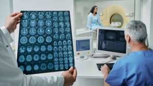 Doctor holding a sheet of brain scans while a technician monitors a patient near an MRI machine.