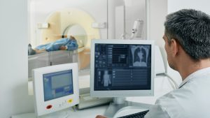 A technician monitors a patient undergoing a CT scan from a computer station, with the scanner visible in the background.