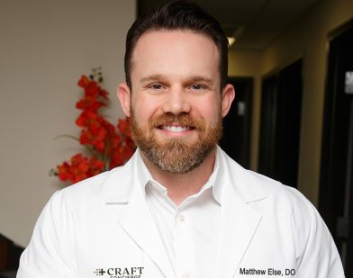 A man with a beard smiling, wearing a white medical coat with name embroidery, standing indoors with red flowers in the background.